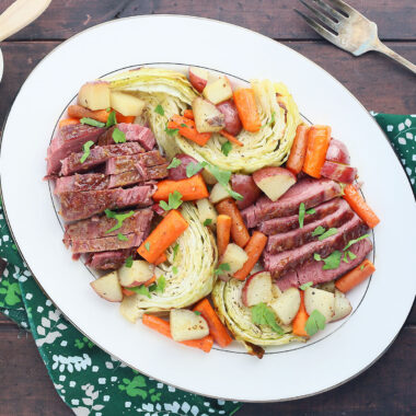 overhead shot of side view of sliced bourbon glazed corned beef and roasted vegetables on a white platter with a green napkin, tarnished silver meat fork, glass of beer and small dish of glaze to the side