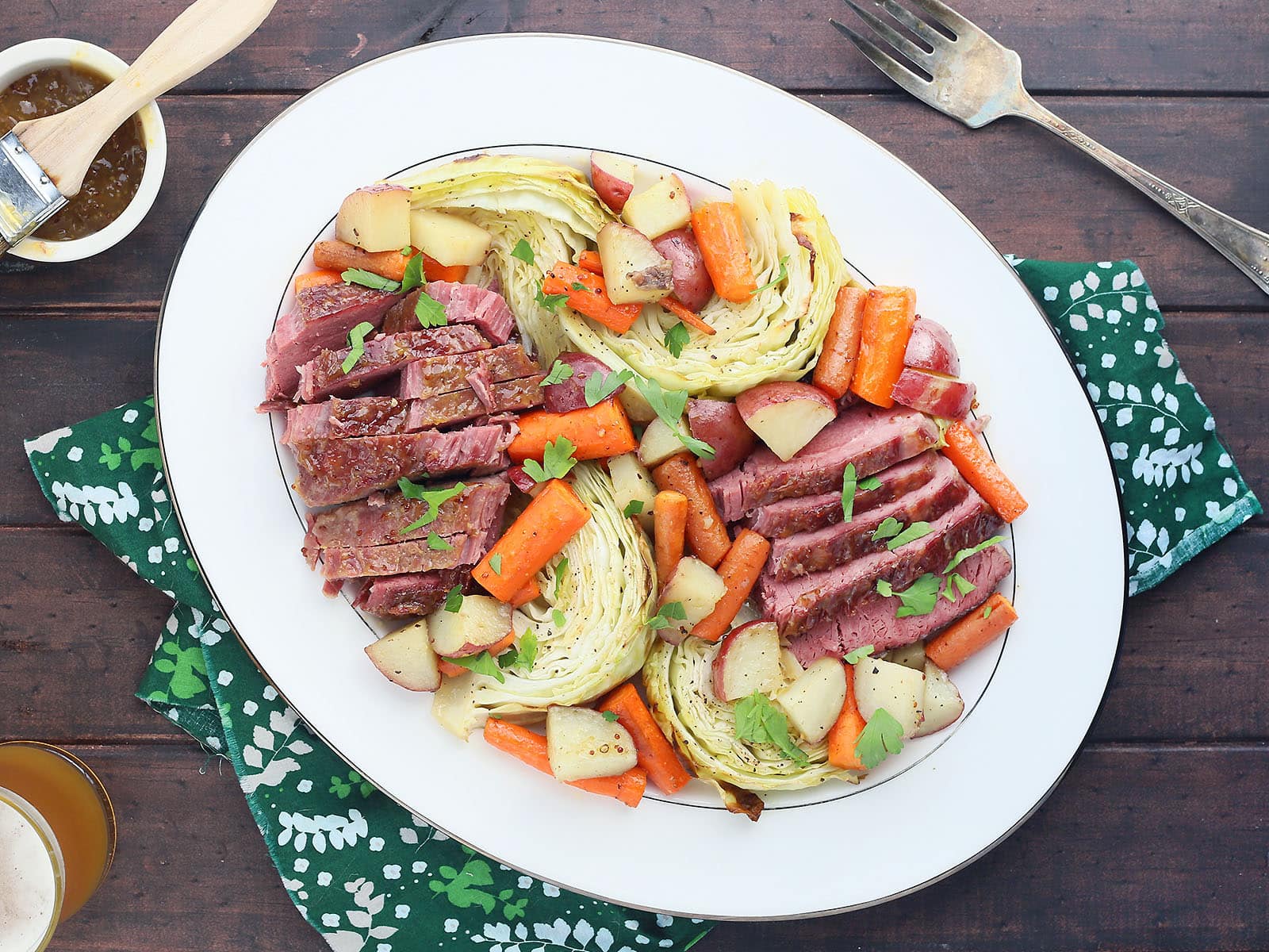 overhead shot of side view of sliced bourbon glazed corned beef and roasted vegetables on a white platter with a green napkin, tarnished silver meat fork, glass of beer and small dish of glaze to the side