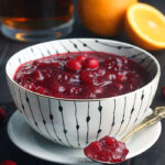 fresh cranberry sauce in a white and black bowl with a spoon of sauce resting on a saucer to the side