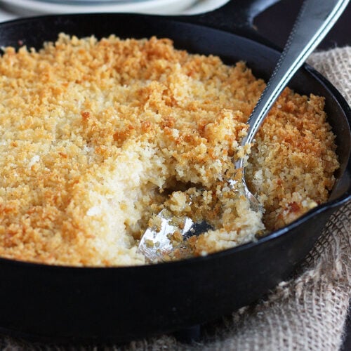 Mashed cauliflower and cheese casserole topped with Panko breadcrumbs in a cast iron skillet with a serving spoon.
