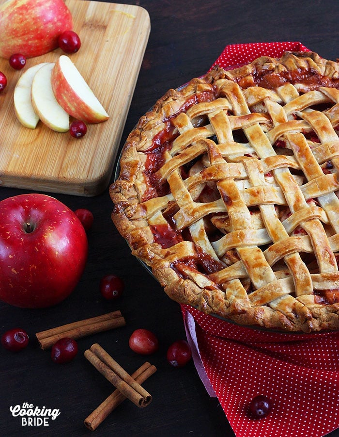 baked cranberry apple pie on a red napkin surrounded by sliced apples, cranberries and cinnamon sticks