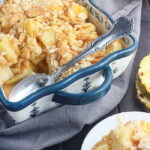 baked pineapple casserole in a blue and white casserole dish with a metal serving spoon and a serving of casserole on the side on a white plate
