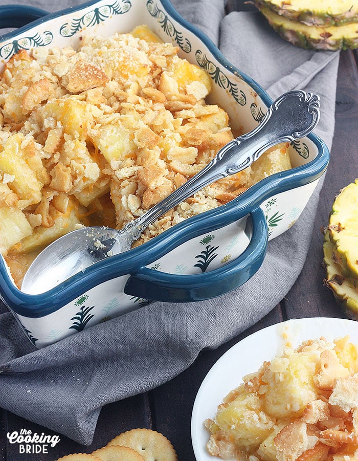 baked pineapple casserole in a blue and white casserole dish with a metal serving spoon and a serving of casserole on the side on a white plate
