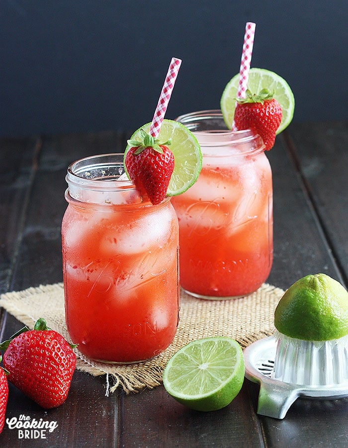 Two glasses of vodka lemonade on a wooden background with strawberries and limes surrounding the glasses 