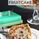 Slice of white fruitcake on a plate with two forks. Fruitcake on a glass pedestal in the background.