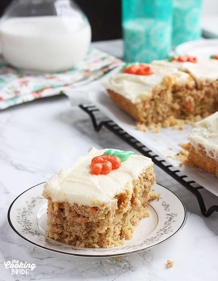 a single slice of carrot cake on a plate with cut cake. glasses and a pitcher of milk in the background