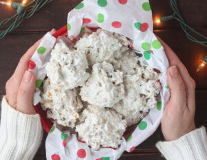 pair of hands holding a container of coconut cornflake cookies on a dark wooden table surrounded by red ornaments and Christmas lights