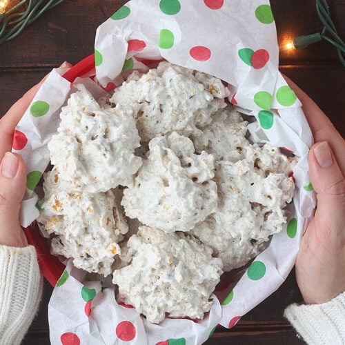 pair of hands holding a container of coconut cornflake cookies on a dark wooden table surrounded by red ornaments and Christmas lights