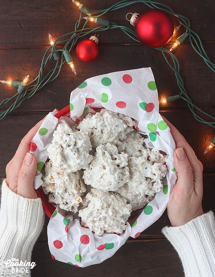 pair of hands holding a container of coconut cornflake cookies on a dark wooden table surrounded by red ornaments and Christmas lights
