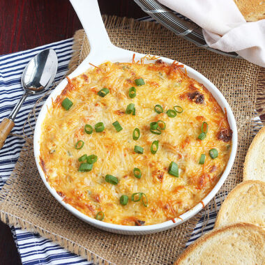 baked crawfish dip in a white ceramic baking dish surrounded by crostini and a serving spoon