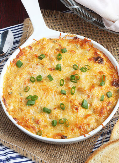 baked crawfish dip in a white ceramic baking dish surrounded by crostini and a serving spoon