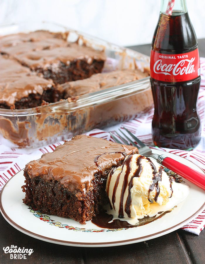 slice of coca cola cake on a white plate with a bottle of soda and the pan of cake in the background