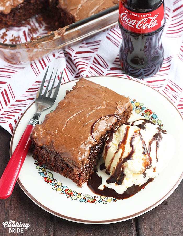 slice of coca cola cake on a white plate with a bottle of soda and the pan of cake in the background