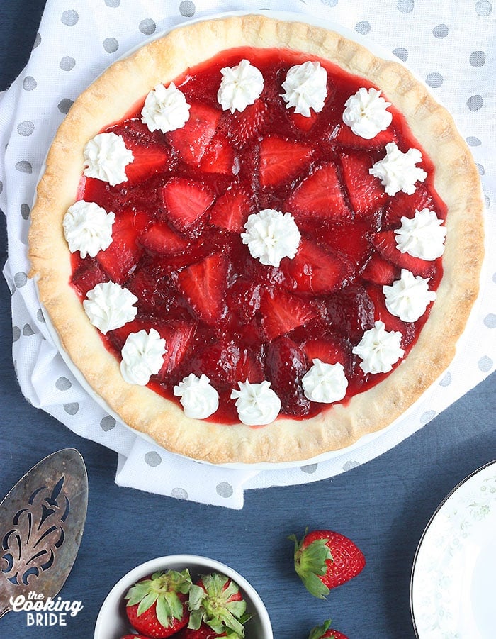 whole strawberry pie on a white and gray polka dotted napkin with a metal cake spatula and a small bowl of strawberries on a blue background
