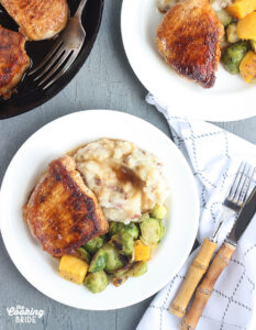 two plated pork chops with mashed potatoes and gravy and roasted vegetables on a white plate