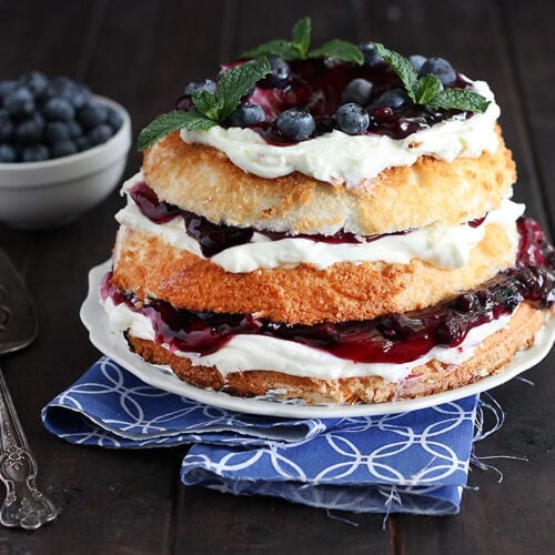 blueberry icebox cake garnished with mint leaves on a white plate served on a blue and white napkin with a silver cake server and a small bowl of blueberries in the background