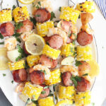 shrimp kabobs with sausage, corn and potatoes threaded on metal skewers on a white platter with a blue and white striped napkin underneath