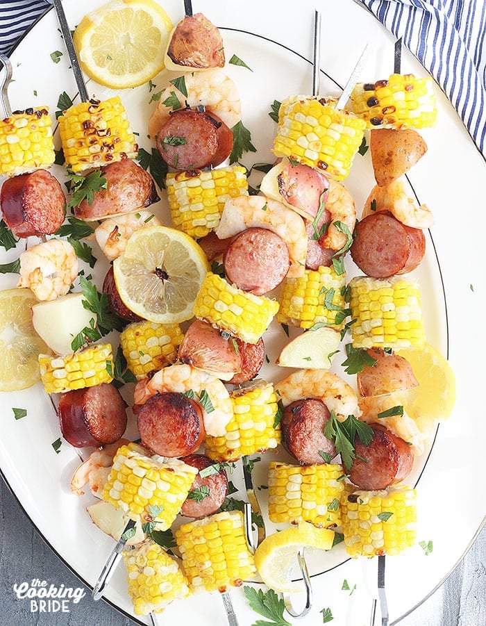 shrimp kabobs with sausage, corn and potatoes threaded on metal skewers on a white platter with a blue and white striped napkin underneath
