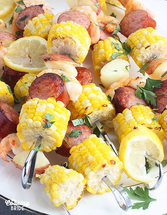 shrimp kabobs with sausage, corn and potatoes threaded on metal skewers on a white platter
