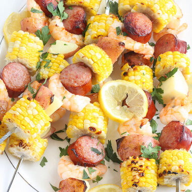 shrimp kabobs with sausage, corn and potatoes threaded on metal skewers on a white platter