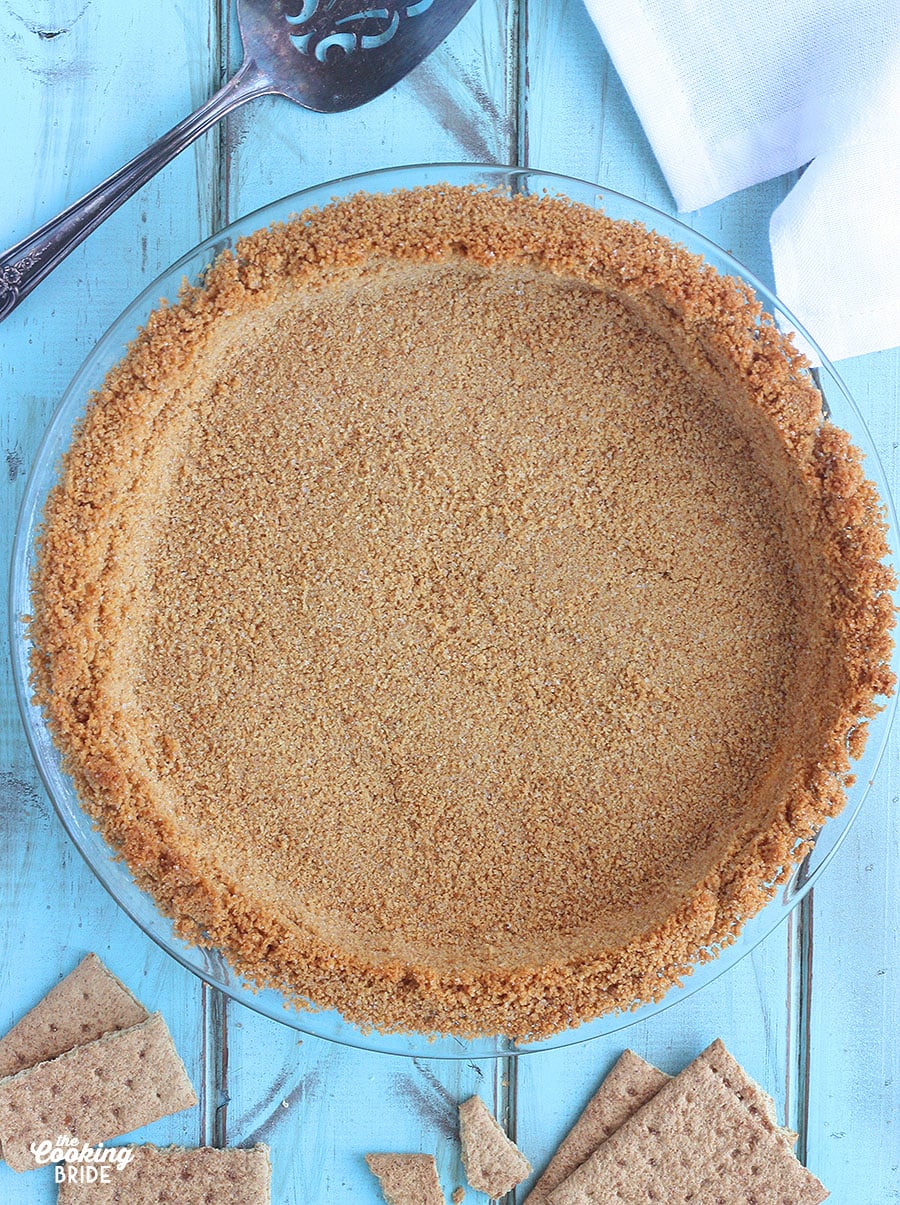 baked graham cracker pie crust against a blue wooden background with a pie serve and graham crackers to the side