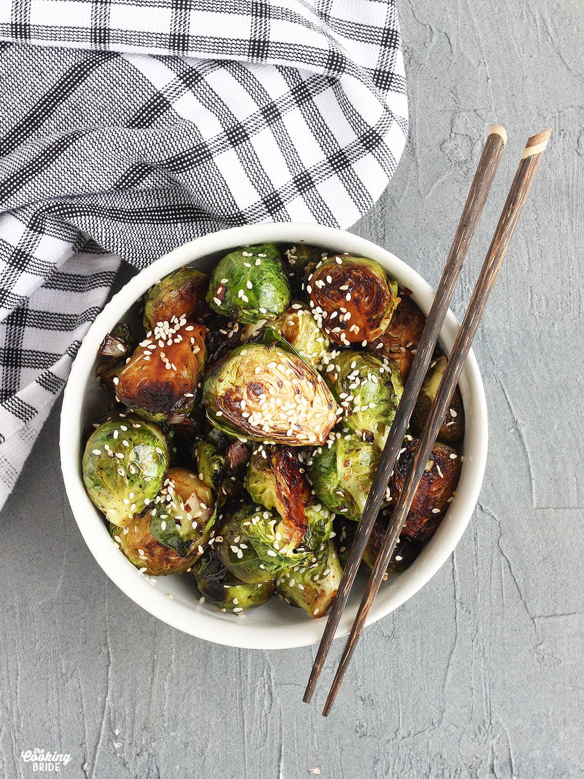 Overhead shot of Stir fried Brussels sprouts garnished with sesame seeds in a white bowl with wooden chopsticks resting on the side of the bowl.