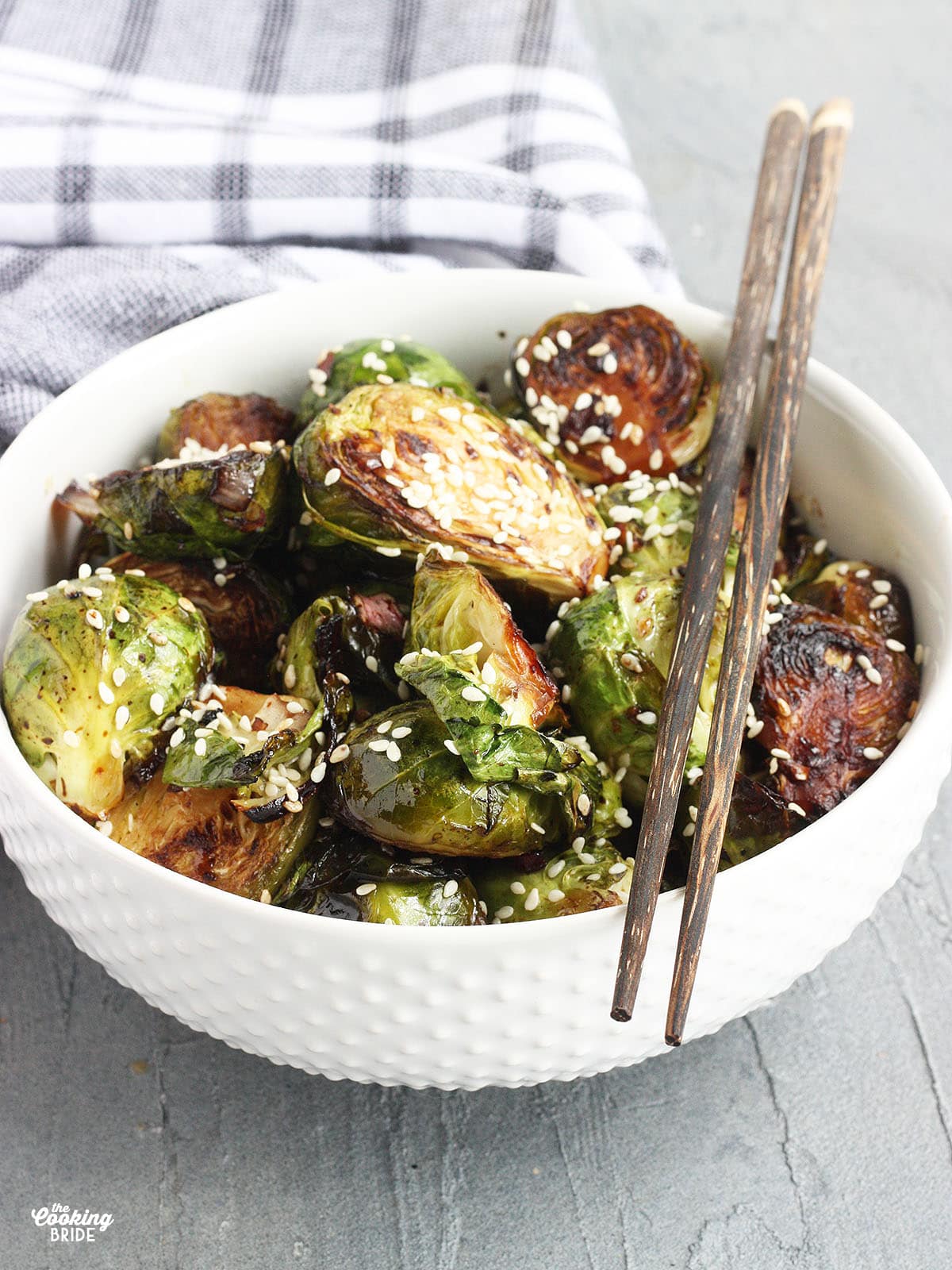 Stir fried Brussels sprouts garnished with sesame seeds in a white bowl with wooden chopsticks resting on the side of the bowl.