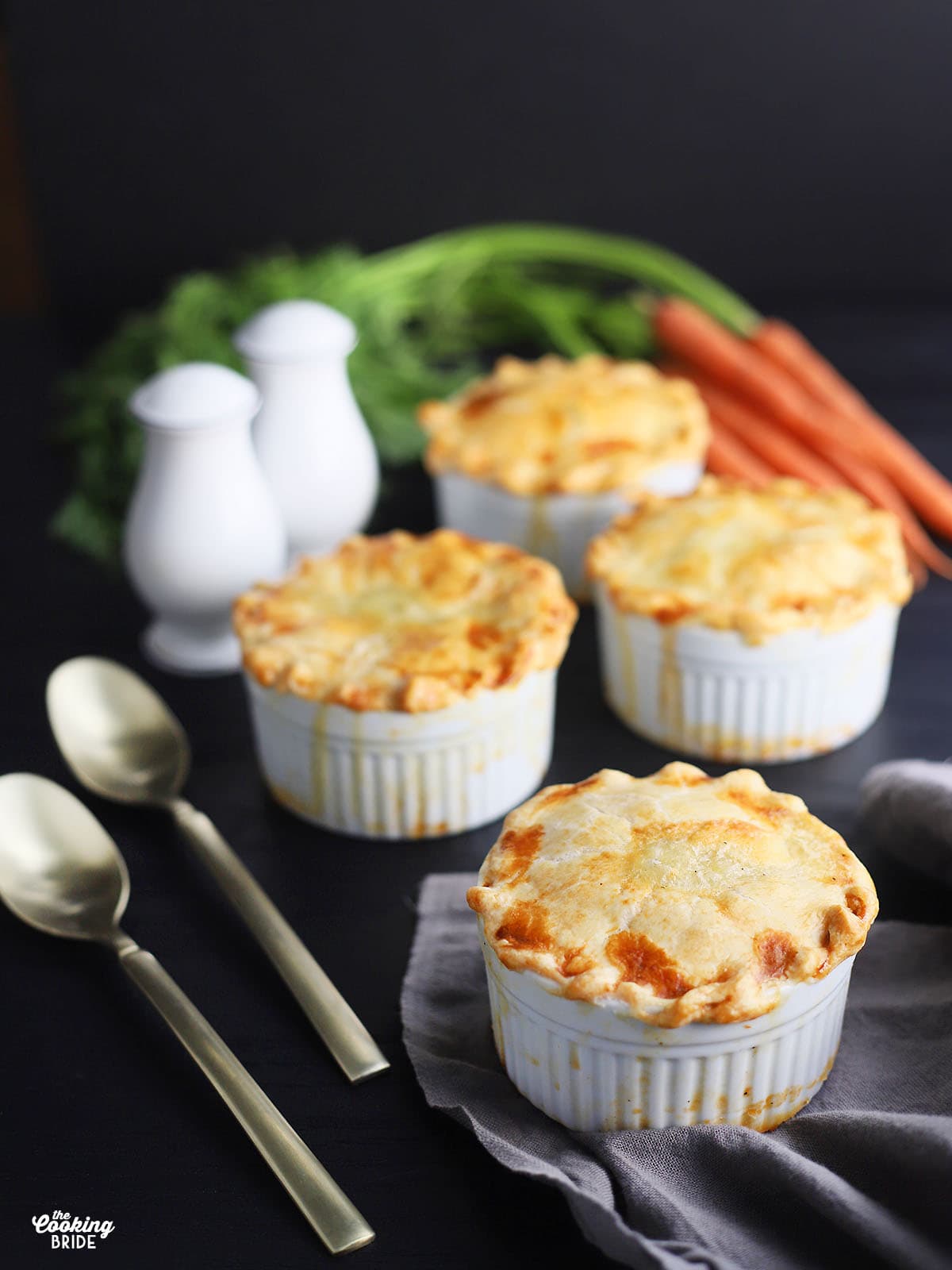 baked mini chicken pot pies on a black background with one bunch of carrots in the background and spoons resting on the side