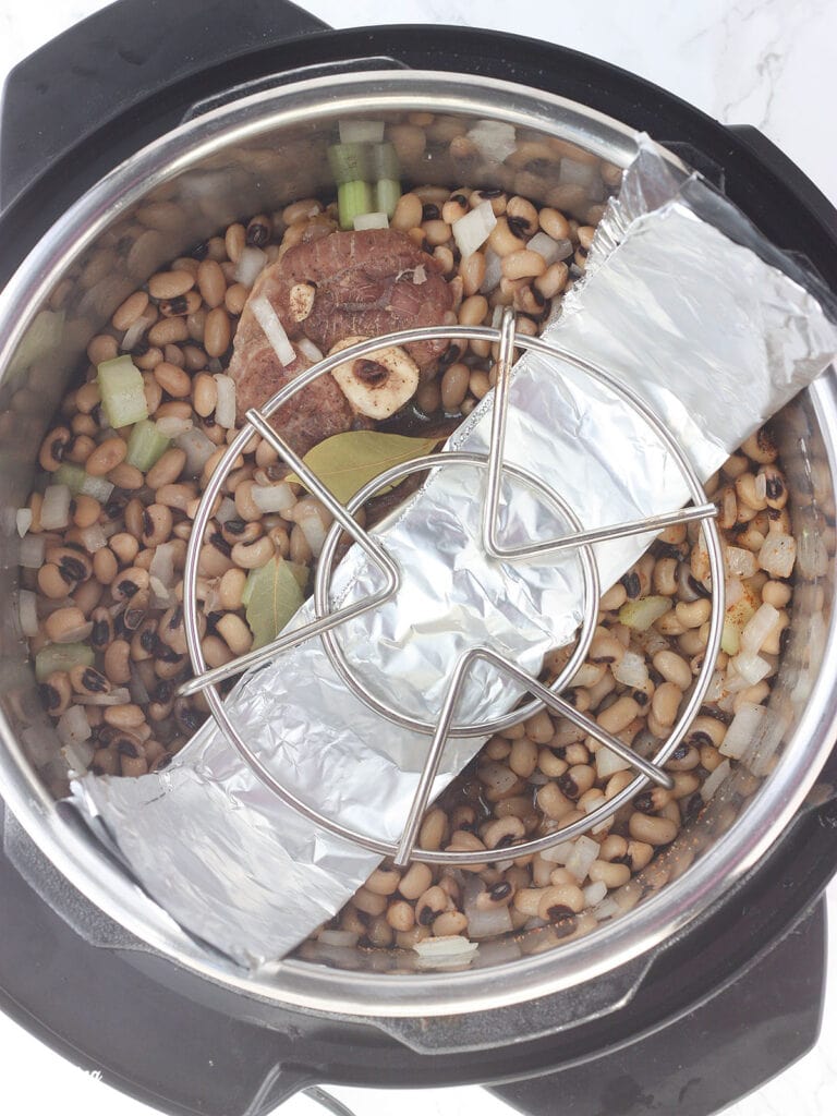 Overhead shot of an Instant Pot with black-eyed peas and ham hock under a trivet and foil sling
