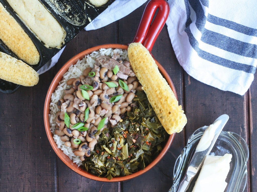 Overhead shot of a red bowl with a handle filled with rice, Hoppin' John and greens. A pan of cornbread and a dish of butter with a knife are to the side.