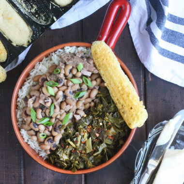 Overhead shot of a red bowl with a handle filled with rice, Hoppin' John and greens. A pan of cornbread and a dish of butter with a knife are to the side.