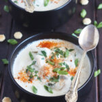 two bowls of oyster stew garnished with green onions and cayenne pepper served in black bowls with a spoon resting on the side