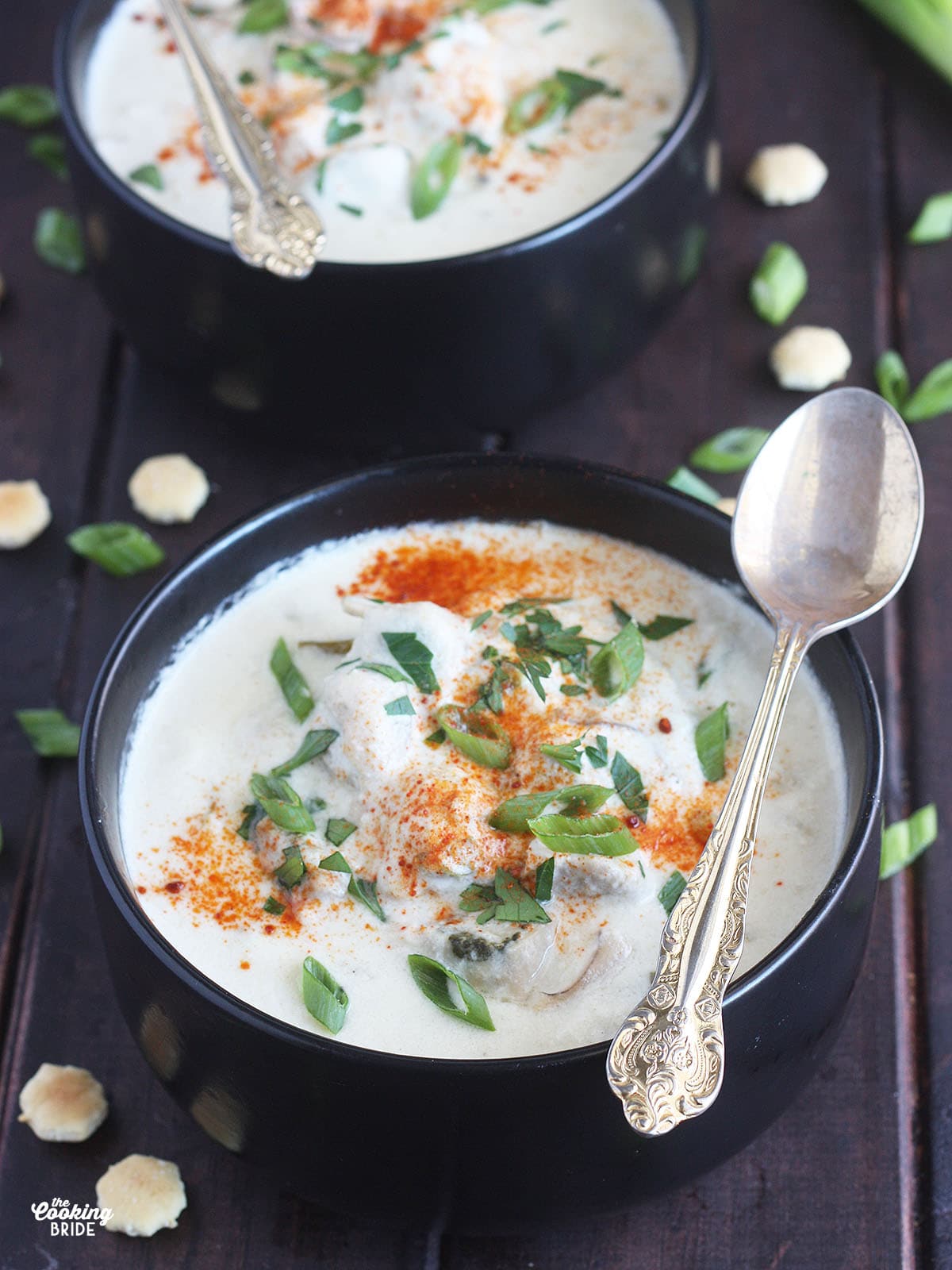 two bowls of oyster stew garnished with green onions and cayenne pepper served in black bowls with a spoon resting on the side