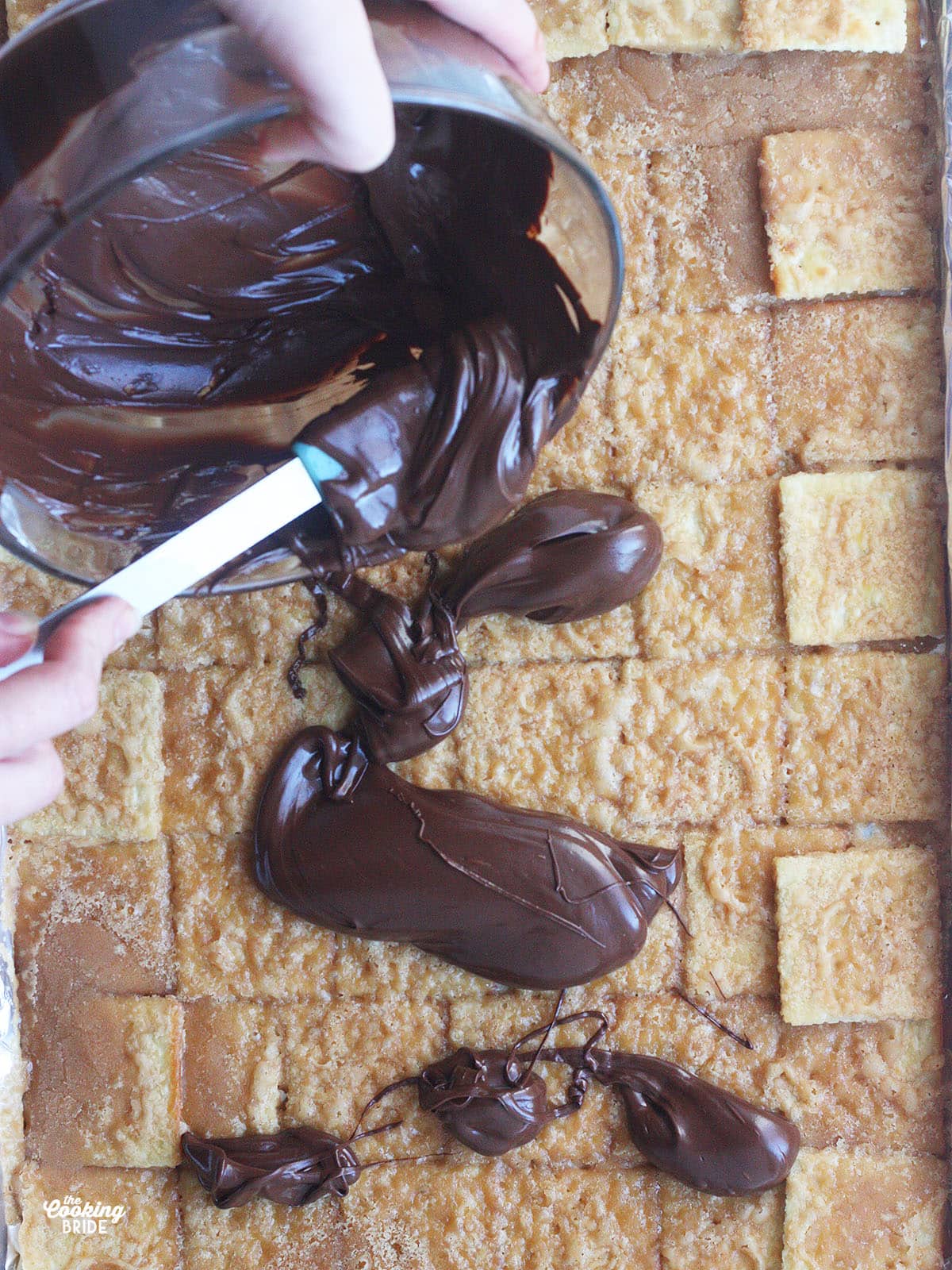 hand pouring melted chocolate from a glass bowl onto the toffee covered saltines