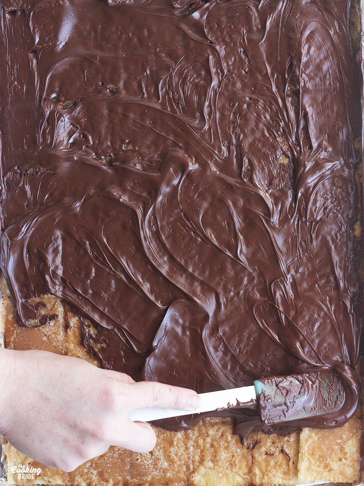 hand using a spatula to spread melted chocolate over the toffee coated crackers