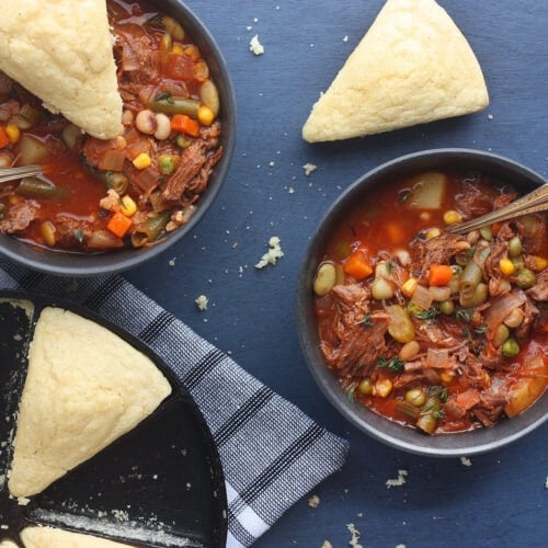 overhead shot of two bowls of vegetable beef soup with two wedges of cornbread and a pan of cornbread to the side