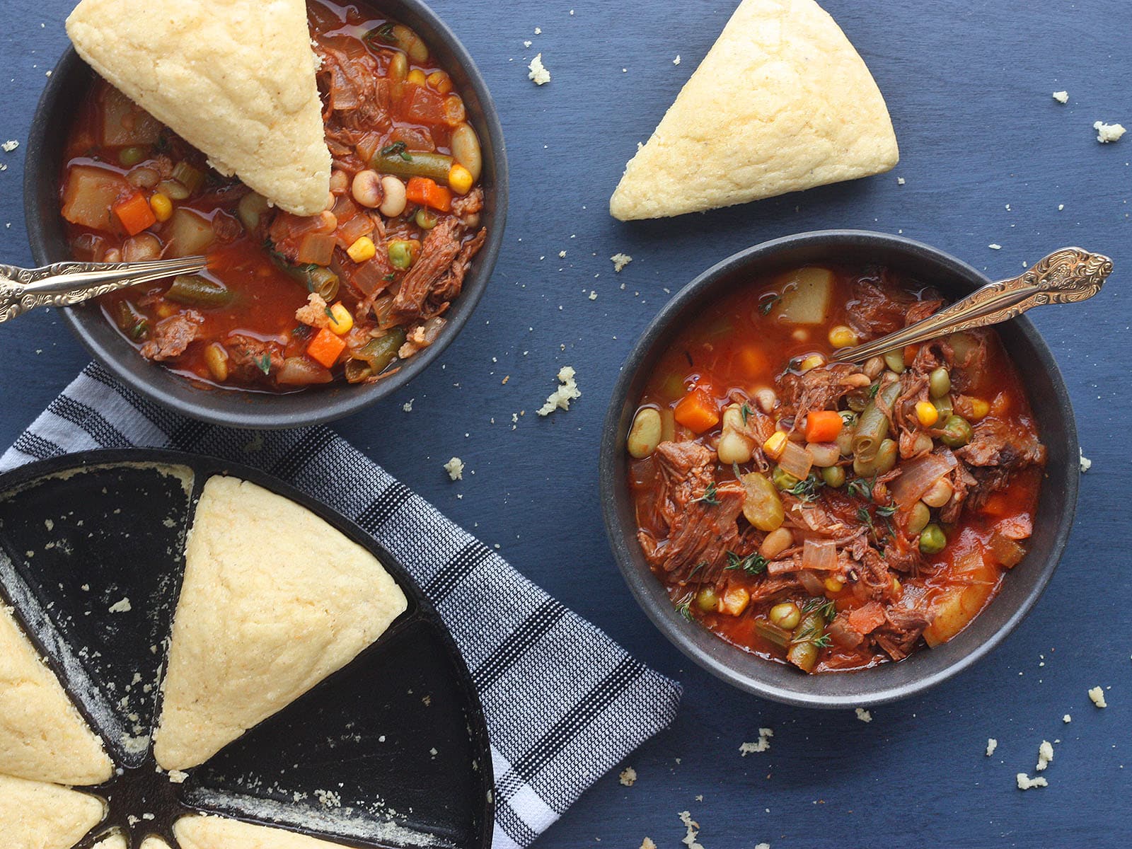 overhead shot of two bowls of vegetable beef soup with two wedges of cornbread and a pan of cornbread to the side