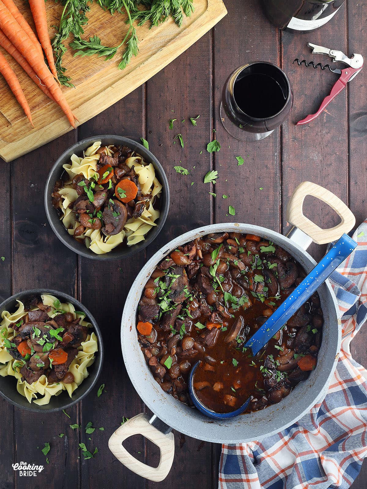 dutch oven full of venison bourguignon with a blue soup ladle, two bowls of venison bourguignon over egg noodles and a glass of red wine to the side