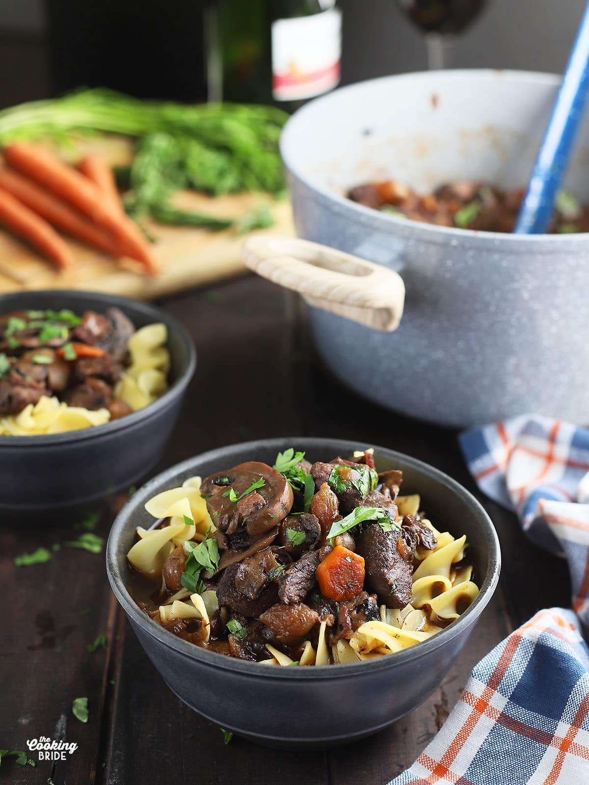 bowl of venison bourguignon over egg noodles with a second bowl, a Dutch oven, carrots, a cutting board and red wine in the background