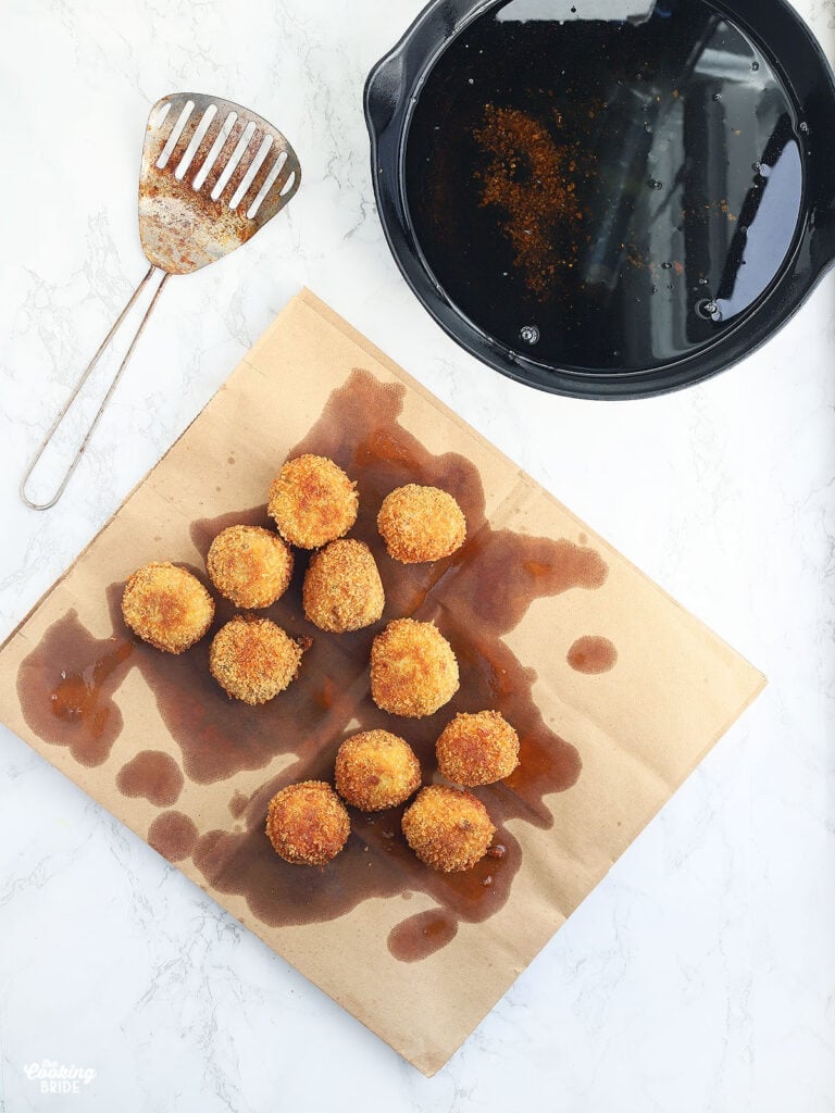 fried boudin balls draining on a brown paper sack with a slotted metal spoon and cast iron skillet of oil to the side