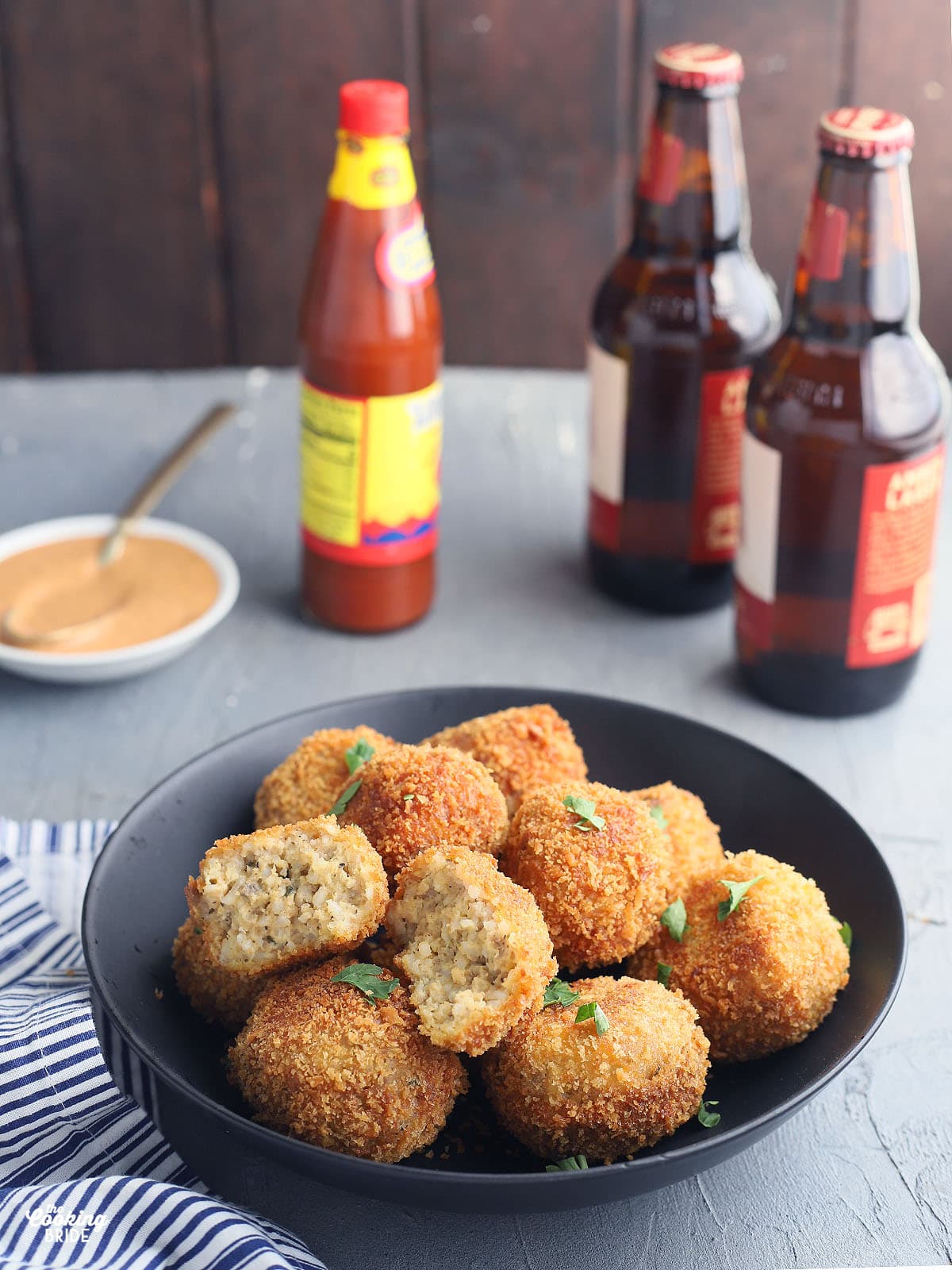 fried boudin balls in a black serving bowl with a small dish of dipping sauce, hot sauce and two bottles of beer in the background