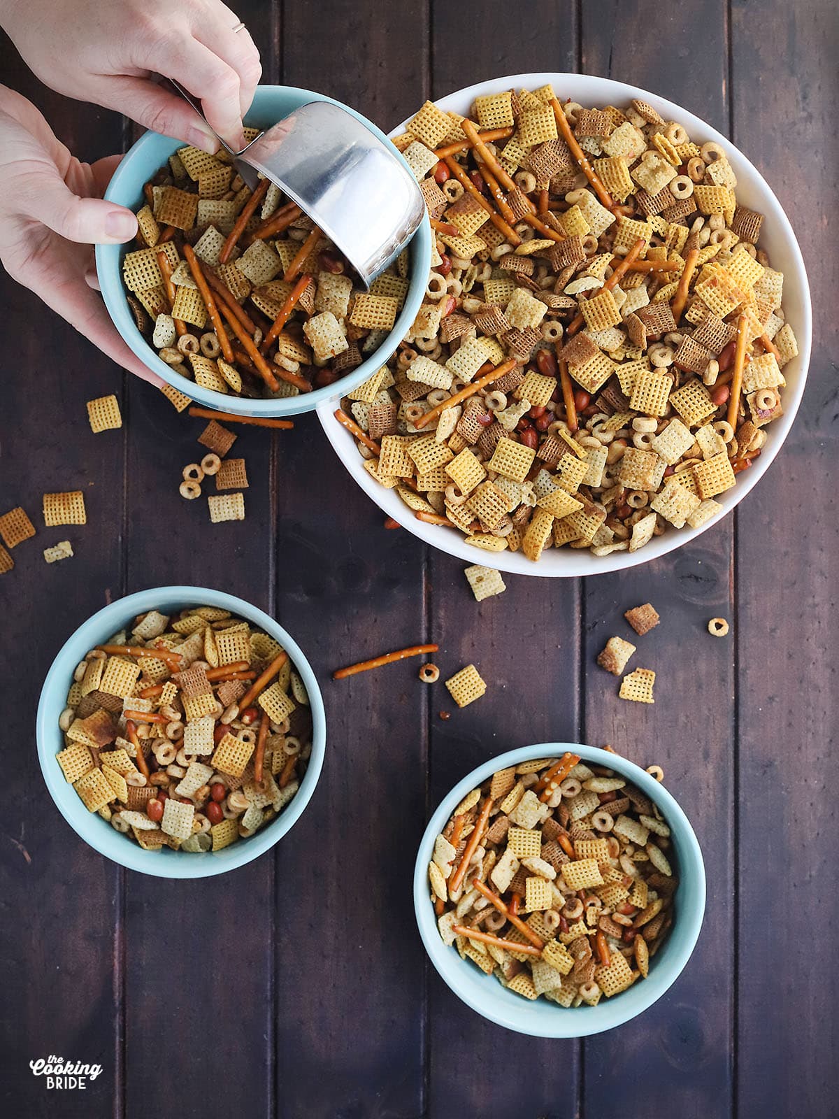 hand using a metal measuring cup to measure out homemade chex mix into blue serving bowls