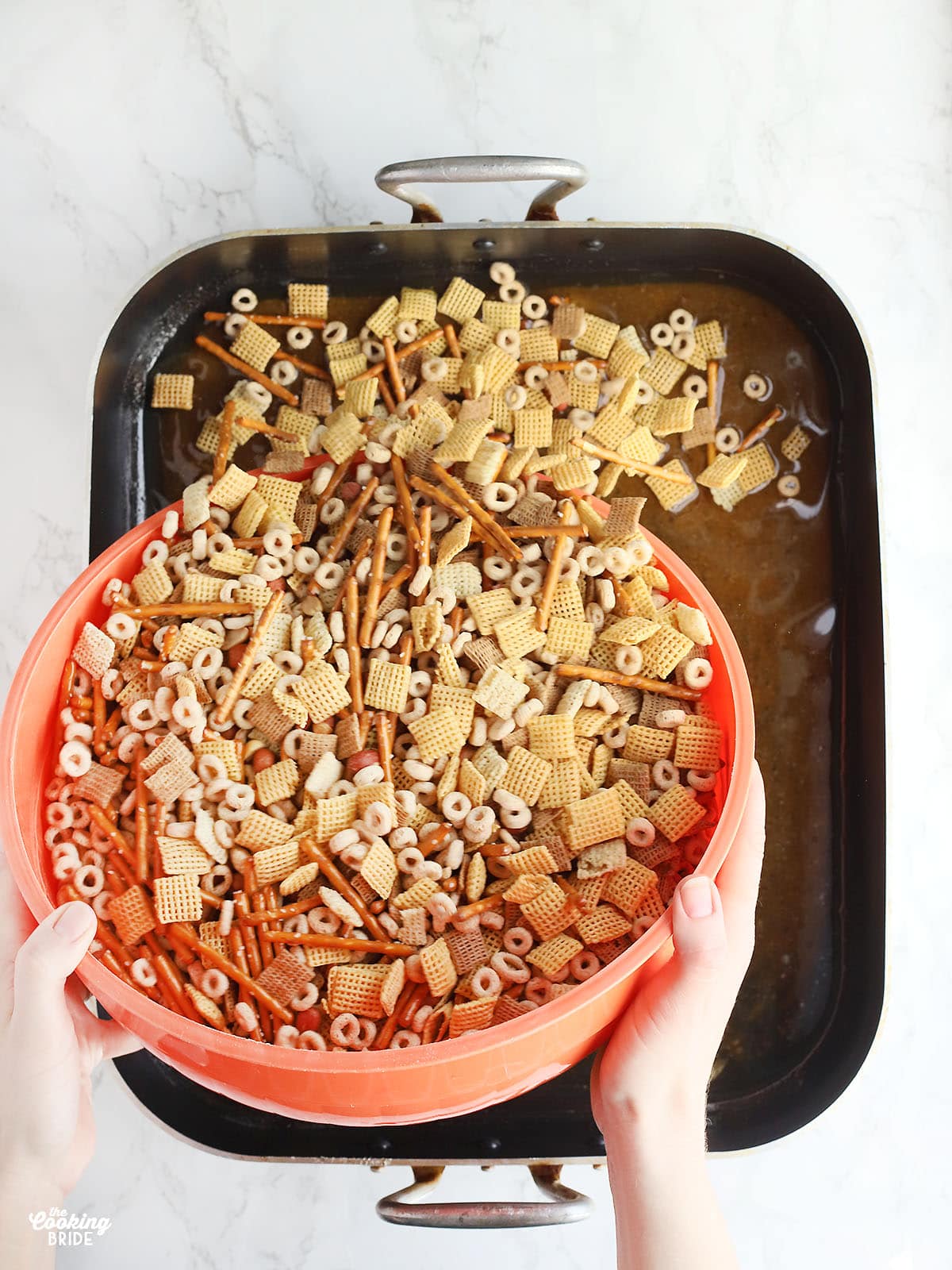 hand pouring an orange bowl of mixed chex mix ingredients into a roasting pan of melted butter and Worcestershire sauce