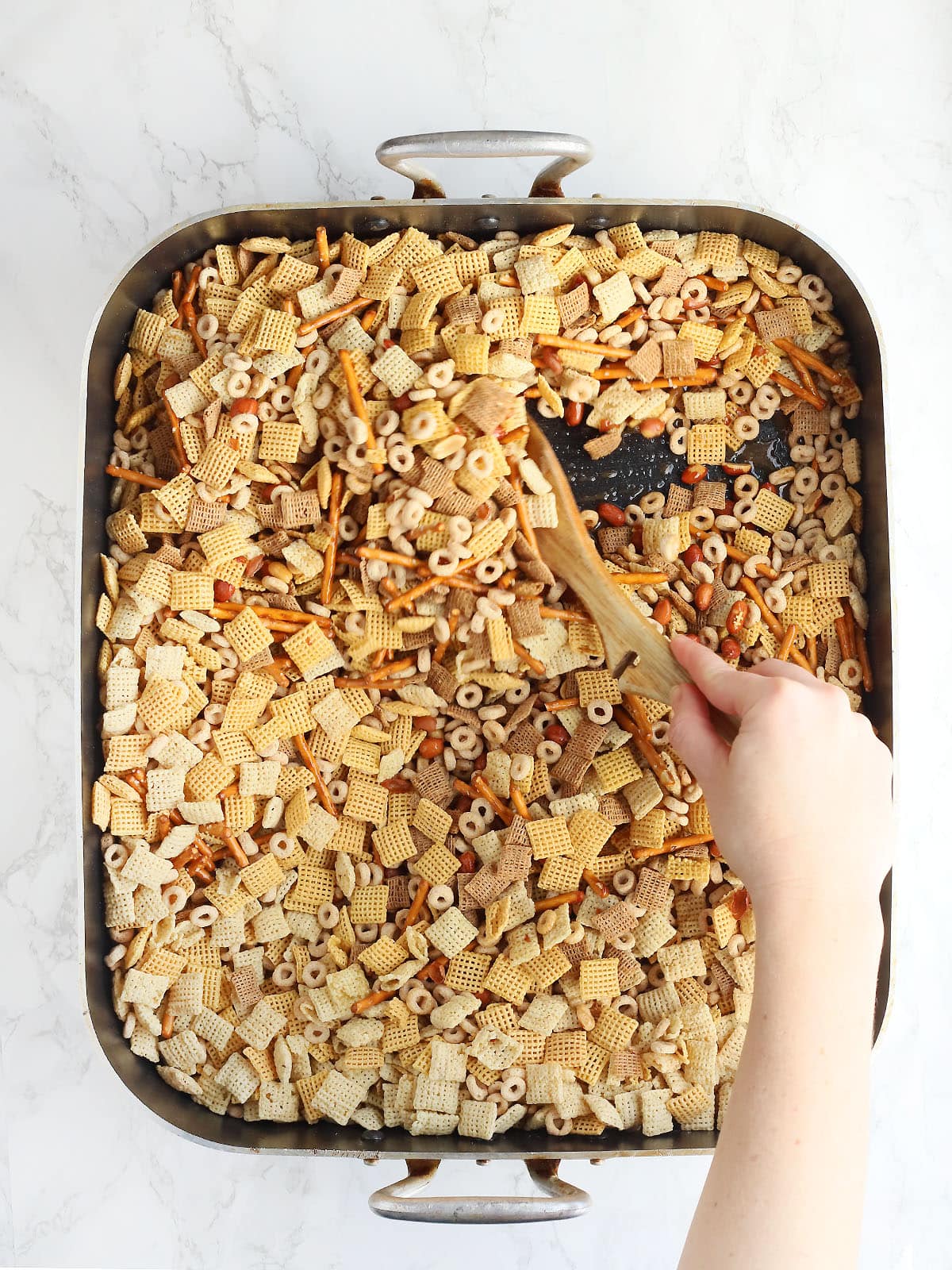 Hand using a wooden spoon to stir Chex mix in a large roasting pan.