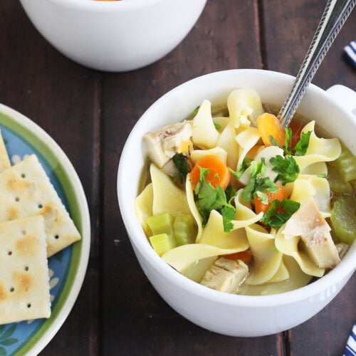 a white cup of chicken noodle soup with a plate of saltines to the side