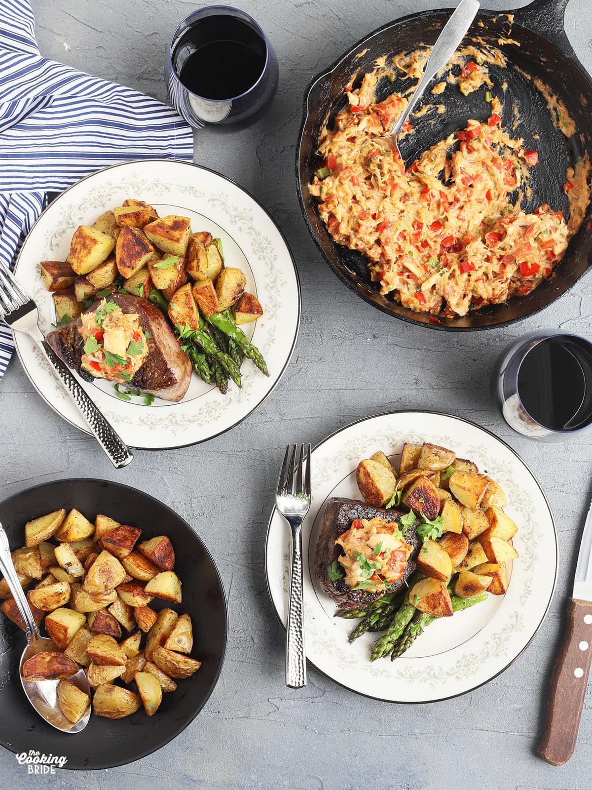 overhead shot of two filets on white China plates garnished with crab topping and served with roasted potatoes and asparagus. A skillet of crab topping, a bowl of roasted potatoes and two glasses of red wine are to the side