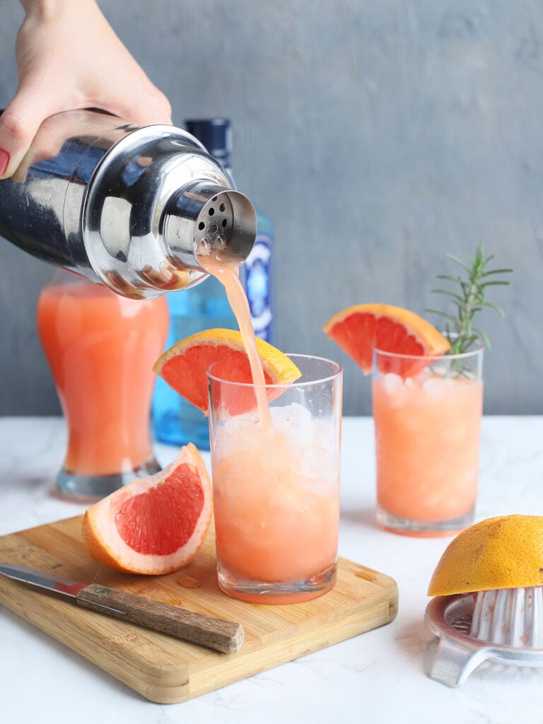 hand pouring a grapefruit cocktail from a shaker into a glass garnished with a grapefruit wedge and a sprig of rosemary