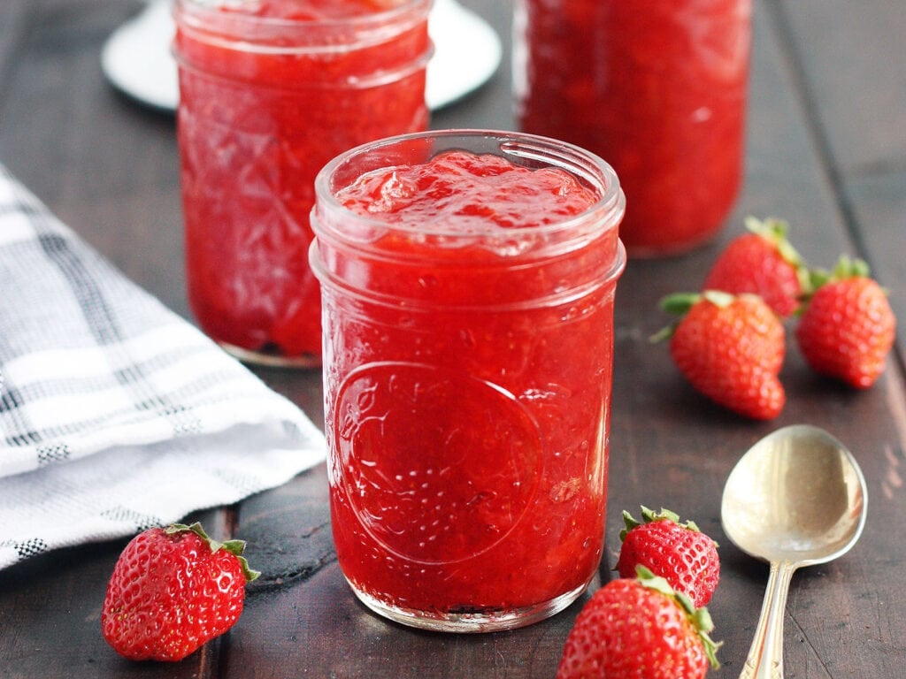 Three jars of homemade strawberry preserves on a wooden table with fresh strawberries and a spoon to the side, colander of fresh strawberries in the background.