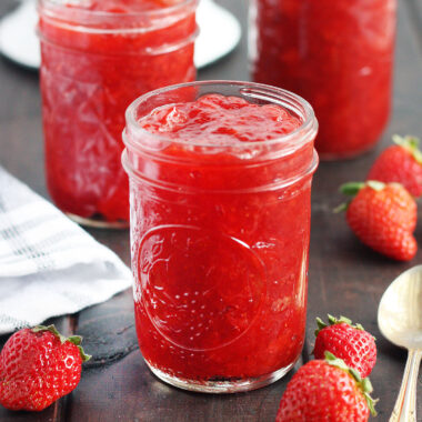 Three jars of homemade strawberry preserves on a wooden table with fresh strawberries and a spoon to the side, colander of fresh strawberries in the background.