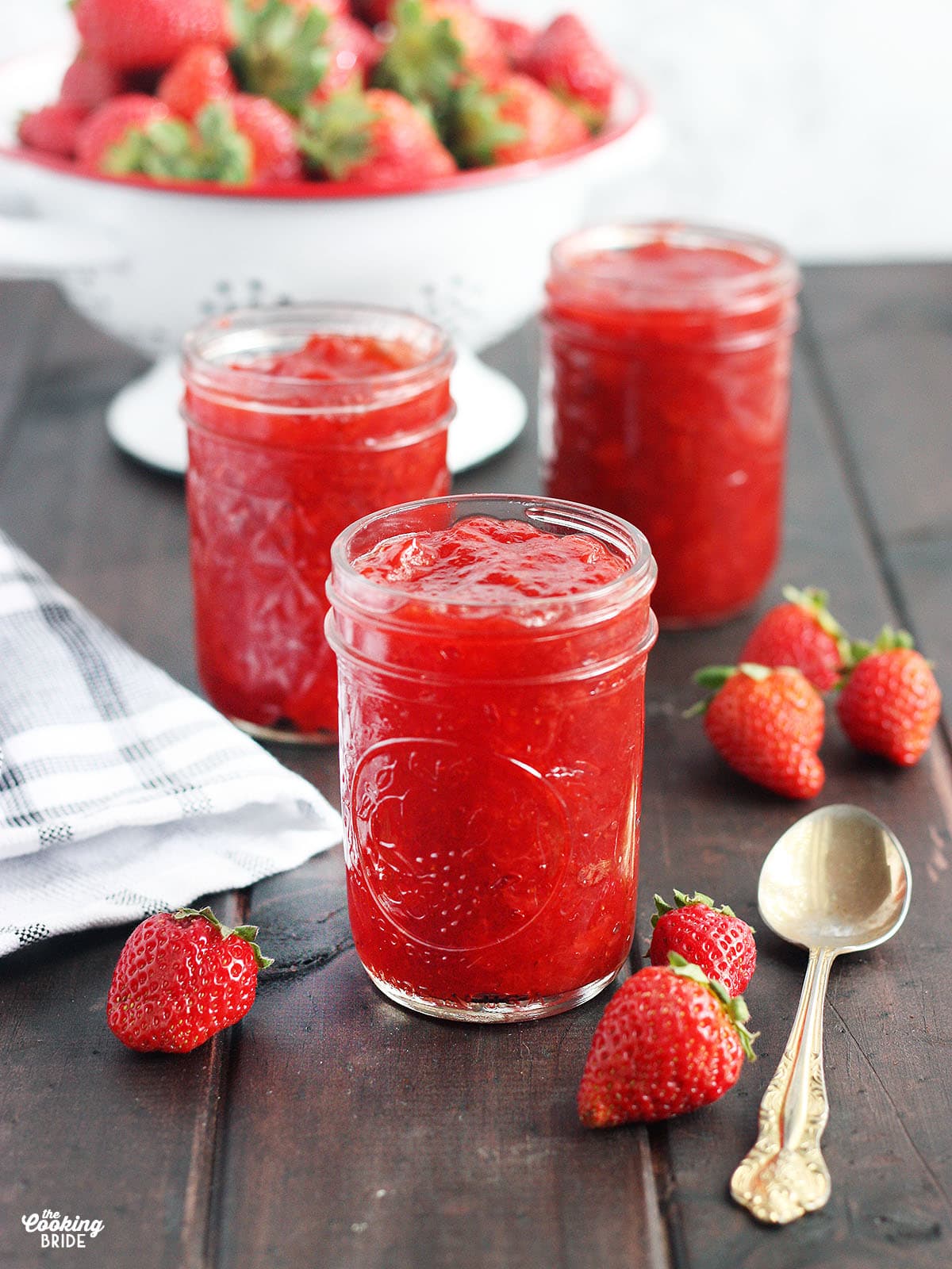 three jars of homemade strawberry preserves on a wooden table with fresh strawberries and a spoon to the side, colander of fresh strawberries in the background